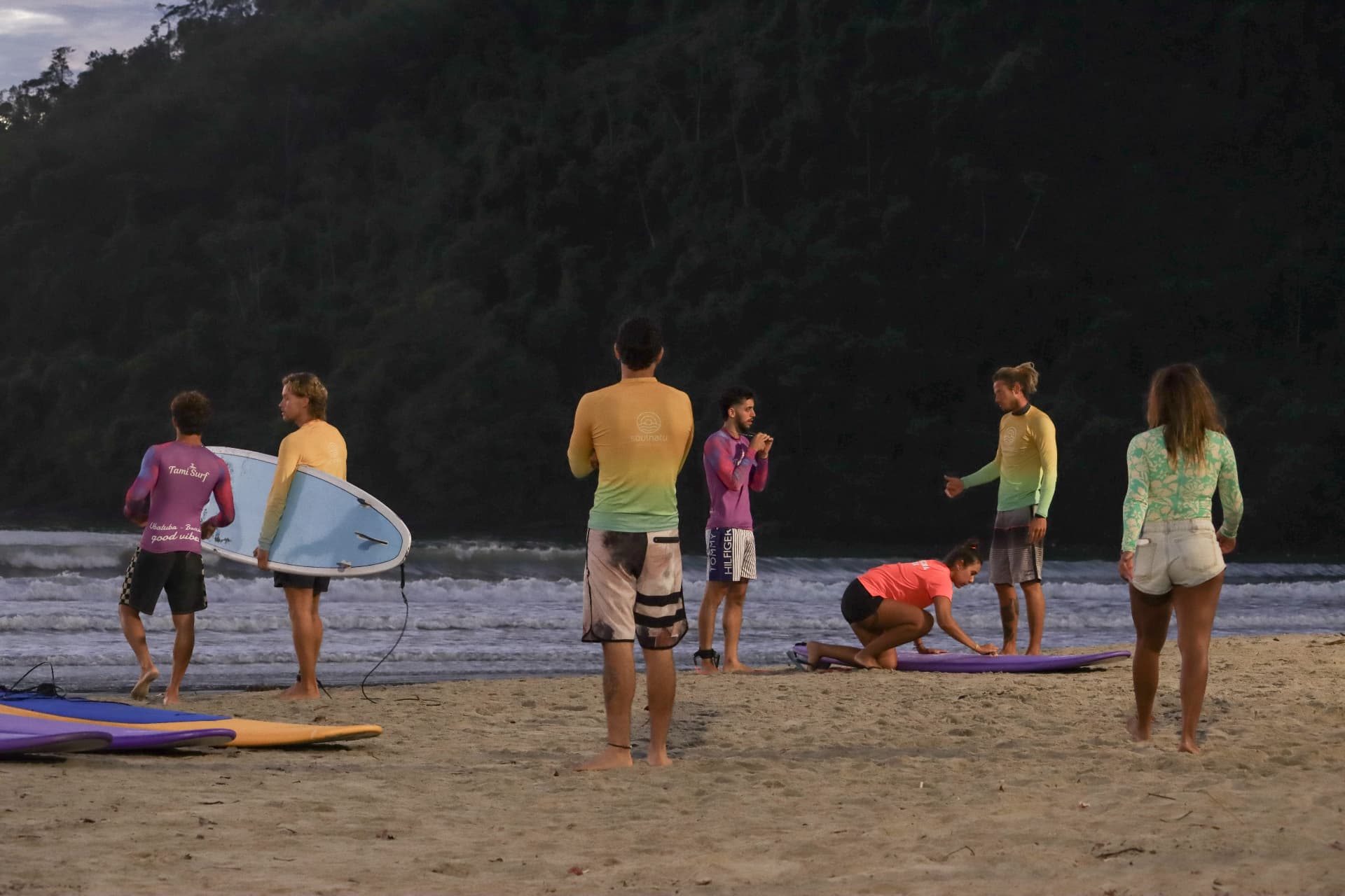 Aula de surf na areia ao entardecer, com pranchas coloridas no areião